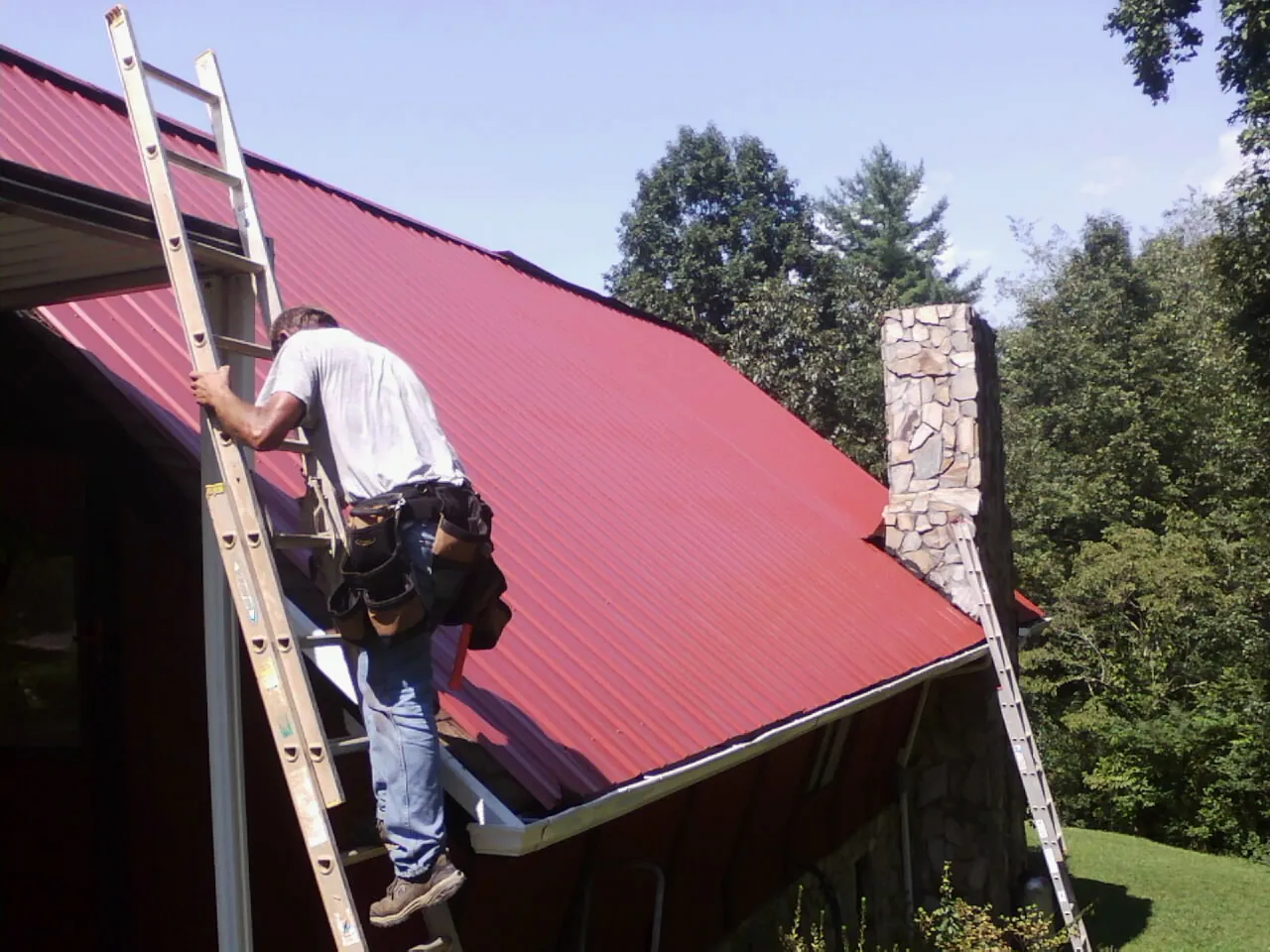 Roofer on a ladder beside a red metal roof and stone chimney — Soffit & Fascia Repair by Roofing Brothers in Luzerne